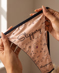 Close-up of the She. Slippery When Wet thong held open to show the full doodle print, Slippery script and black elastic trim